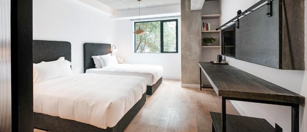 A minimalist twin room with dark headboards, concrete walls and a window onto trees.