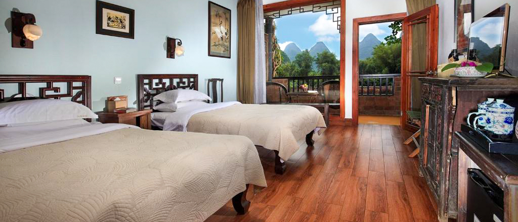 A twin room with carved wooden beds and an open balcony door looking out to limestone peaks.