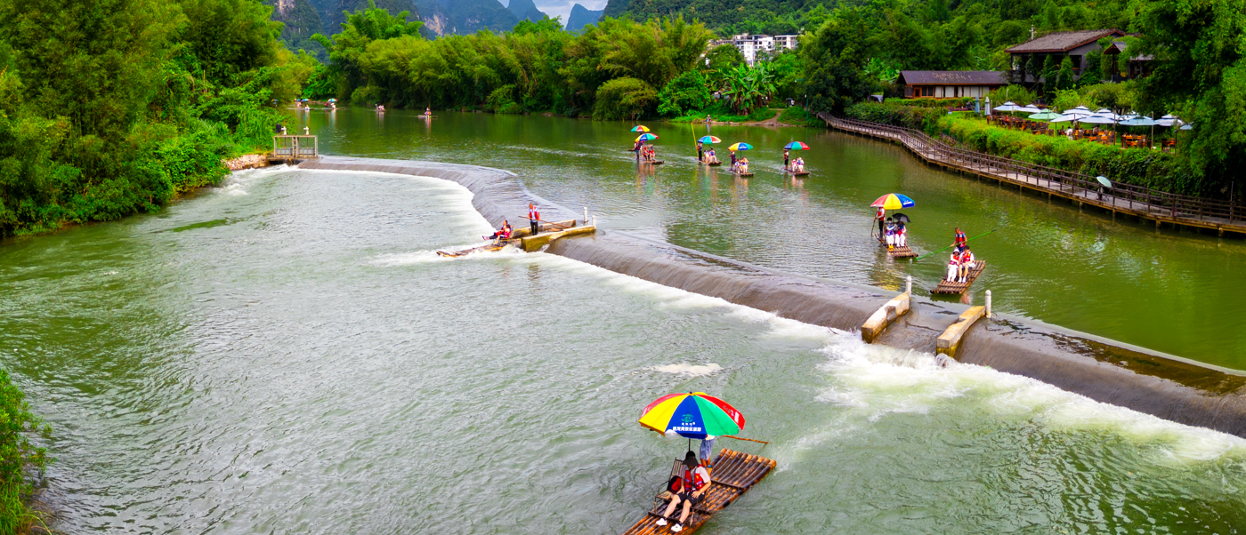 Bamboo rafts drift down a green river between limestone peaks, seen from above.