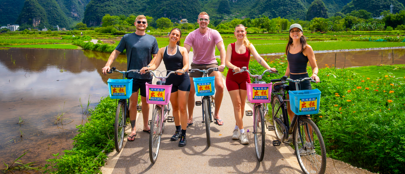 A group of travellers stands with bicycles on a country path beside rice paddies and limestone peaks.