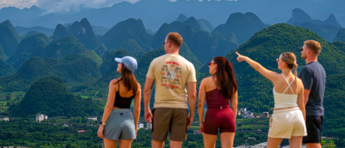Travellers look out from a hilltop over a sea of jagged green limestone peaks under a blue sky.