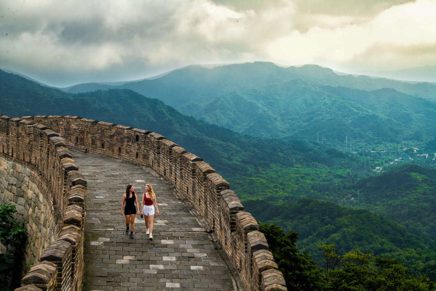 Two travellers walk along the Great Wall as it follows a forested ridge into misty mountains.