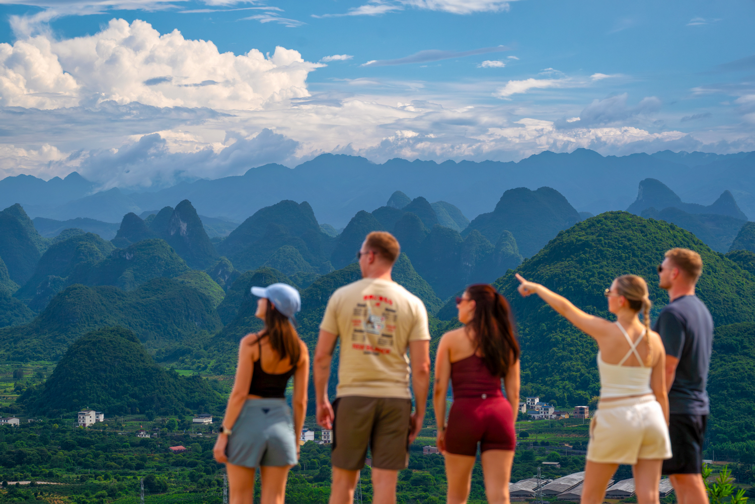 Travellers look out from a hilltop over a sea of jagged green limestone peaks under a blue sky.