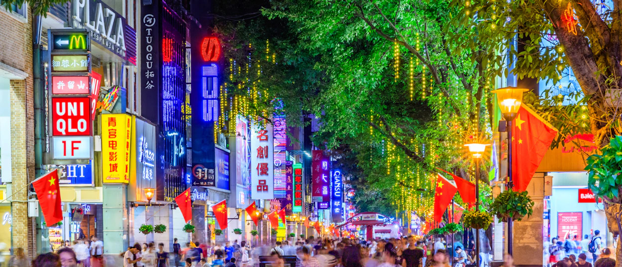 A lively Beijing street at night, bright with colorful neon signs, festive lights, and Chinese flags.