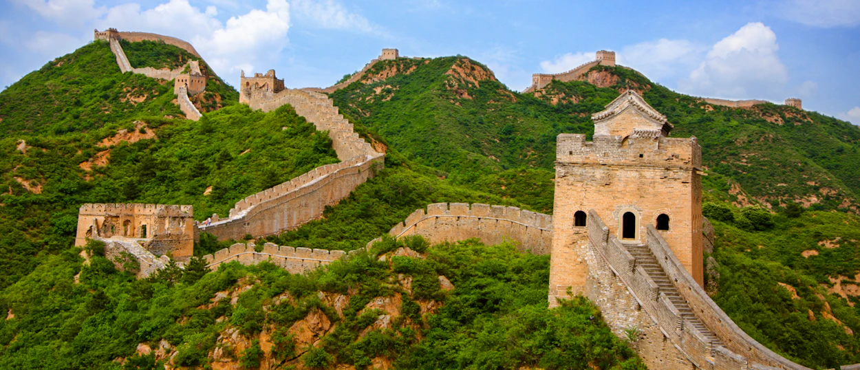 The ancient Great Wall of China with watchtowers winding over green mountains under a blue sky.