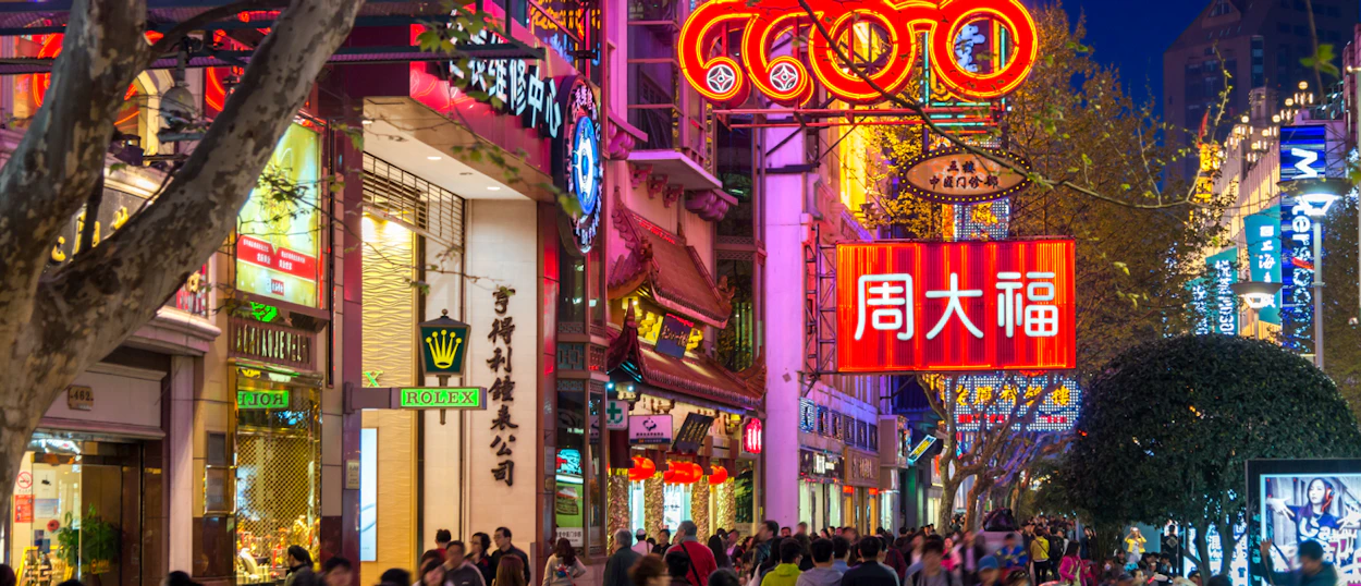 Crowds of people walk along a vibrant Shanghai street at night, illuminated by colorful neon signs.
