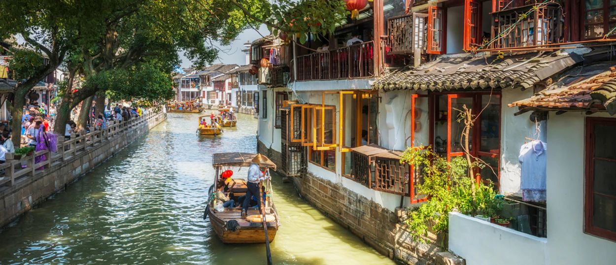 Boats with tourists on a lively canal flowing past traditional buildings and green trees in Shanghai.