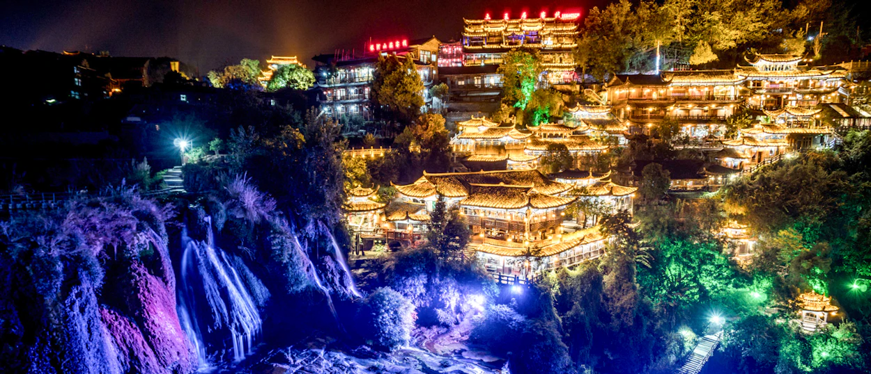 Furong Ancient Town glows at night with colorful lights illuminating the waterfall and traditional buildings.