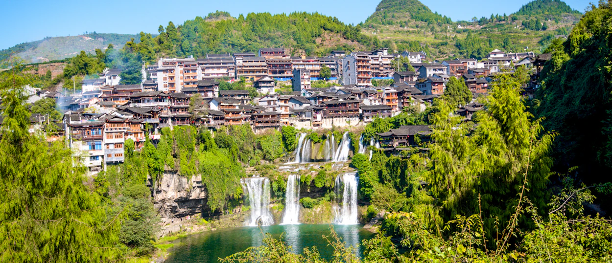 Traditional buildings of Furong Ancient Town overlook multiple waterfalls flowing into a clear pool.