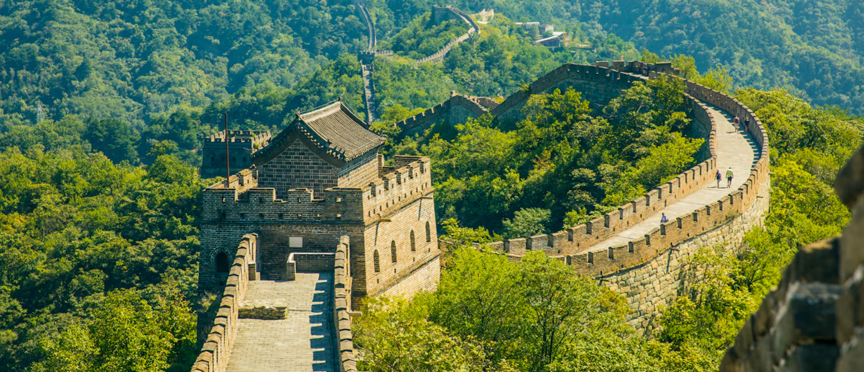 Tourists walk along the ancient Great Wall of China, winding through lush green mountains.
