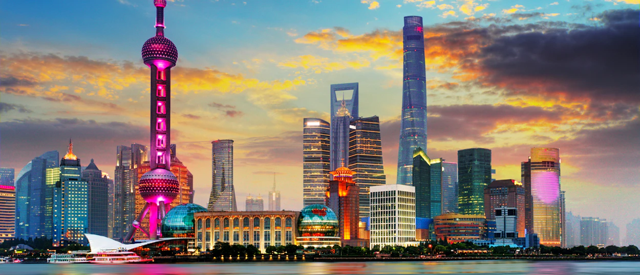 Shanghai's illuminated skyline at dusk, with the Oriental Pearl TV Tower and other skyscrapers reflecting in the river.