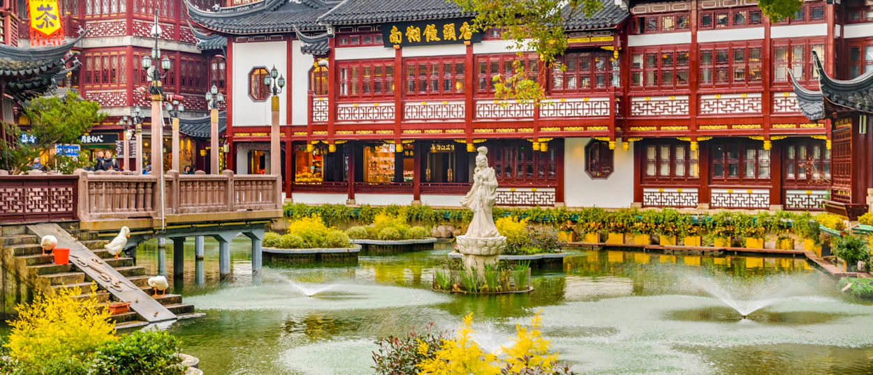 Ornate Chinese garden with traditional red buildings, a serene pond, fountains, and lush greenery in Shanghai.