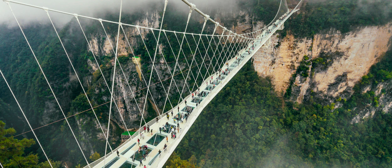 People walk across the Zhangjiajie Glass Bridge, suspended high above a misty, forested valley.