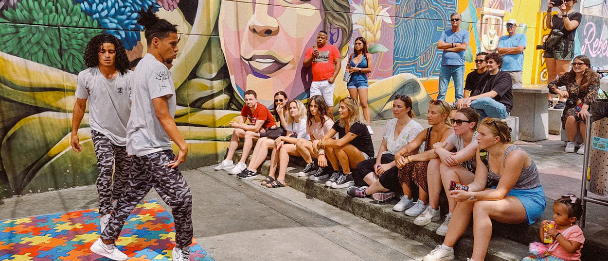Two men perform street dance in front of a vibrant mural as tourists watch in Medellín.