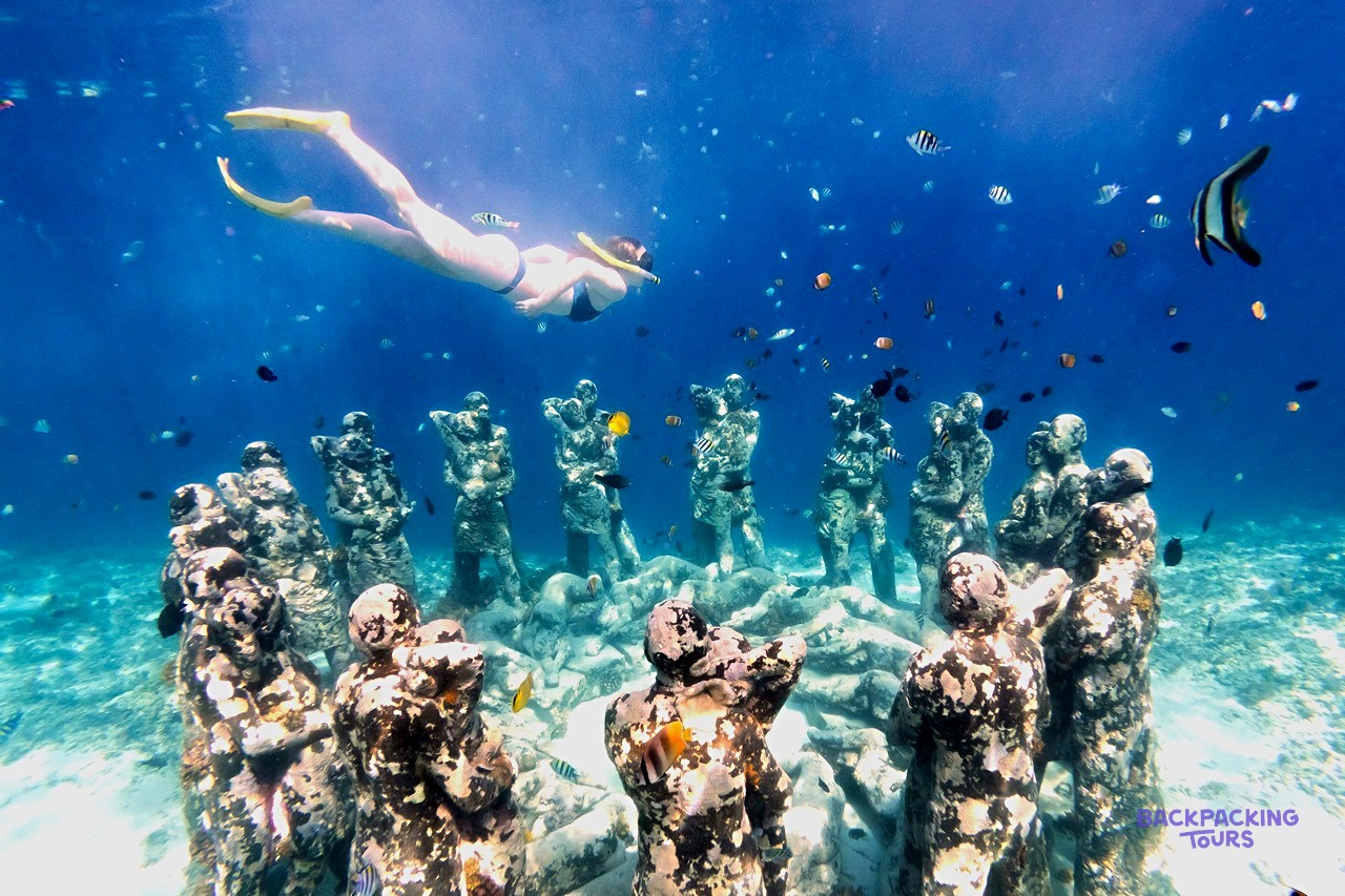 A snorkeller swims above a circle of human-like statues on the seabed, surrounded by colourful fish.