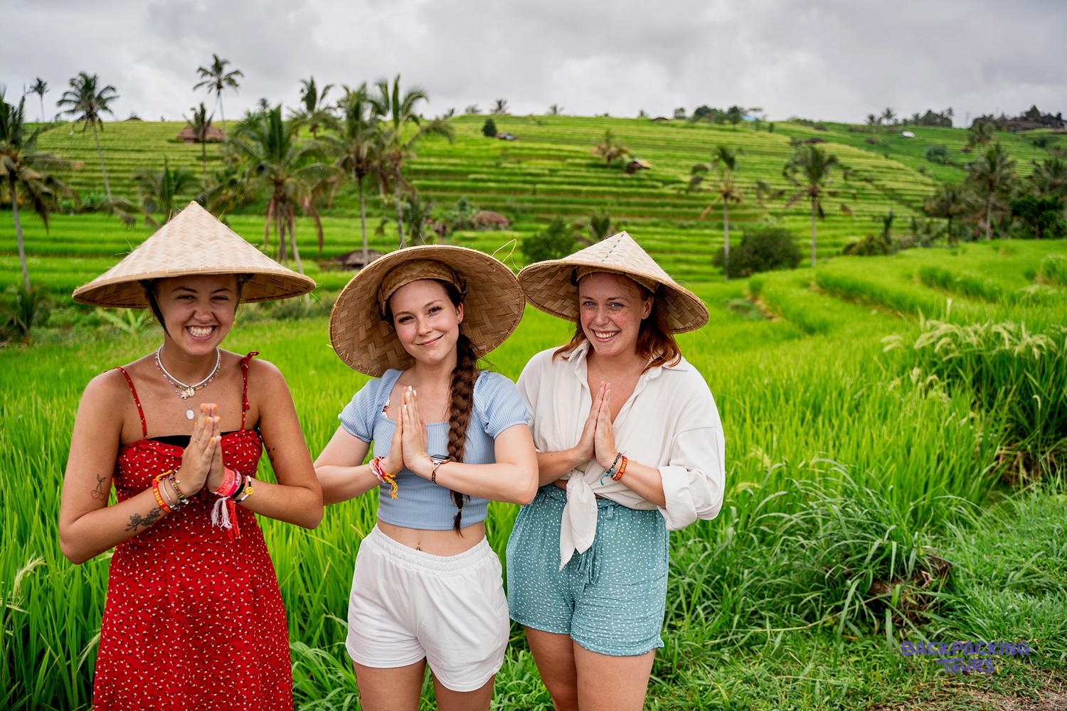 Three young women in straw hats stand with hands pressed together, surrounded by lush green rice terraces and palm trees.