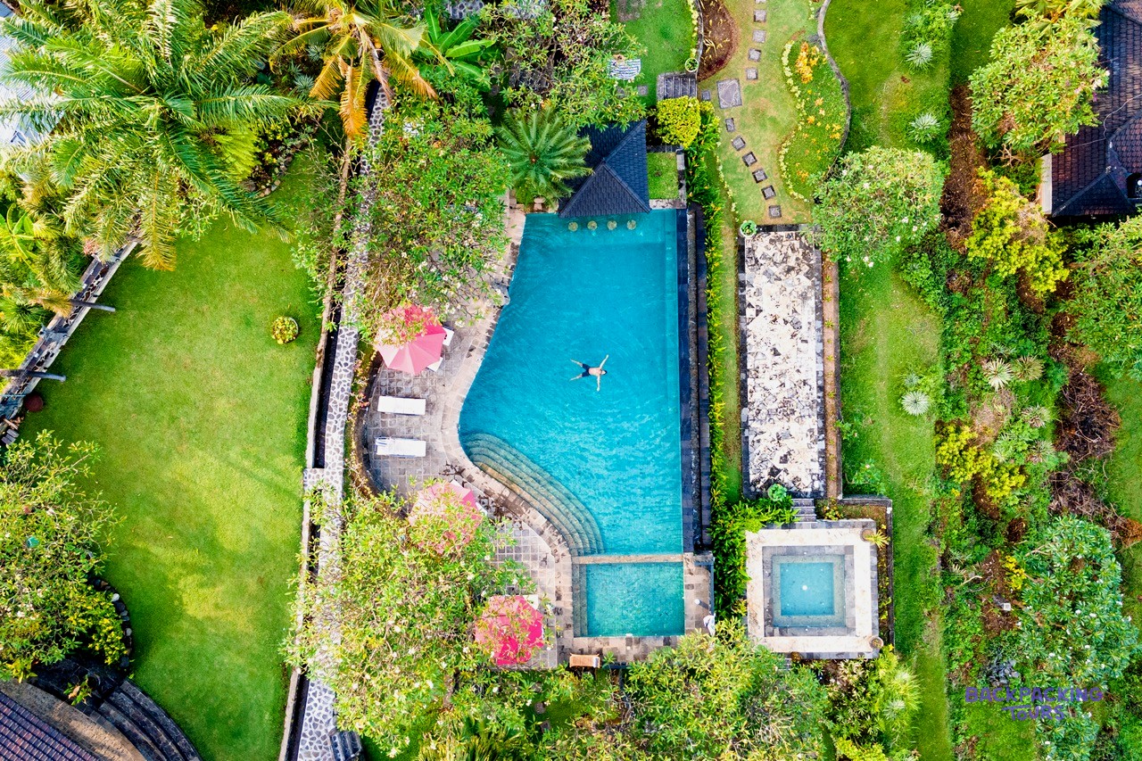 An aerial view shows a person floating in a turquoise villa pool surrounded by lush green gardens and sun loungers.