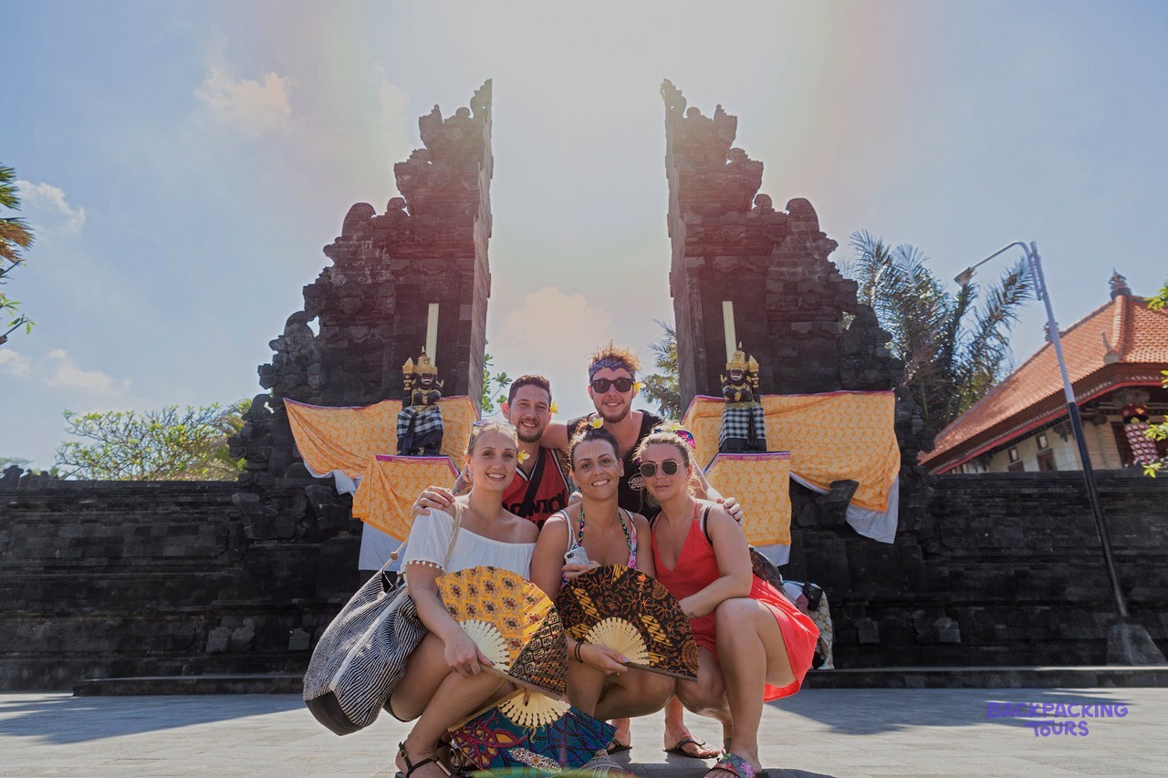 Five young people pose smiling with decorative fans in front of a traditional stone temple gate.