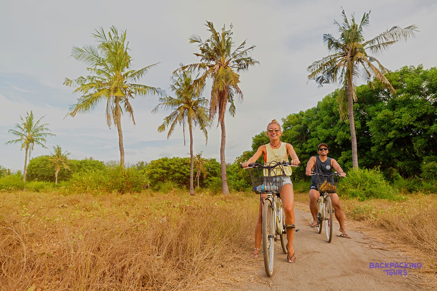 Two people cycle along a dirt path lined with tall palm trees and dry golden grass.