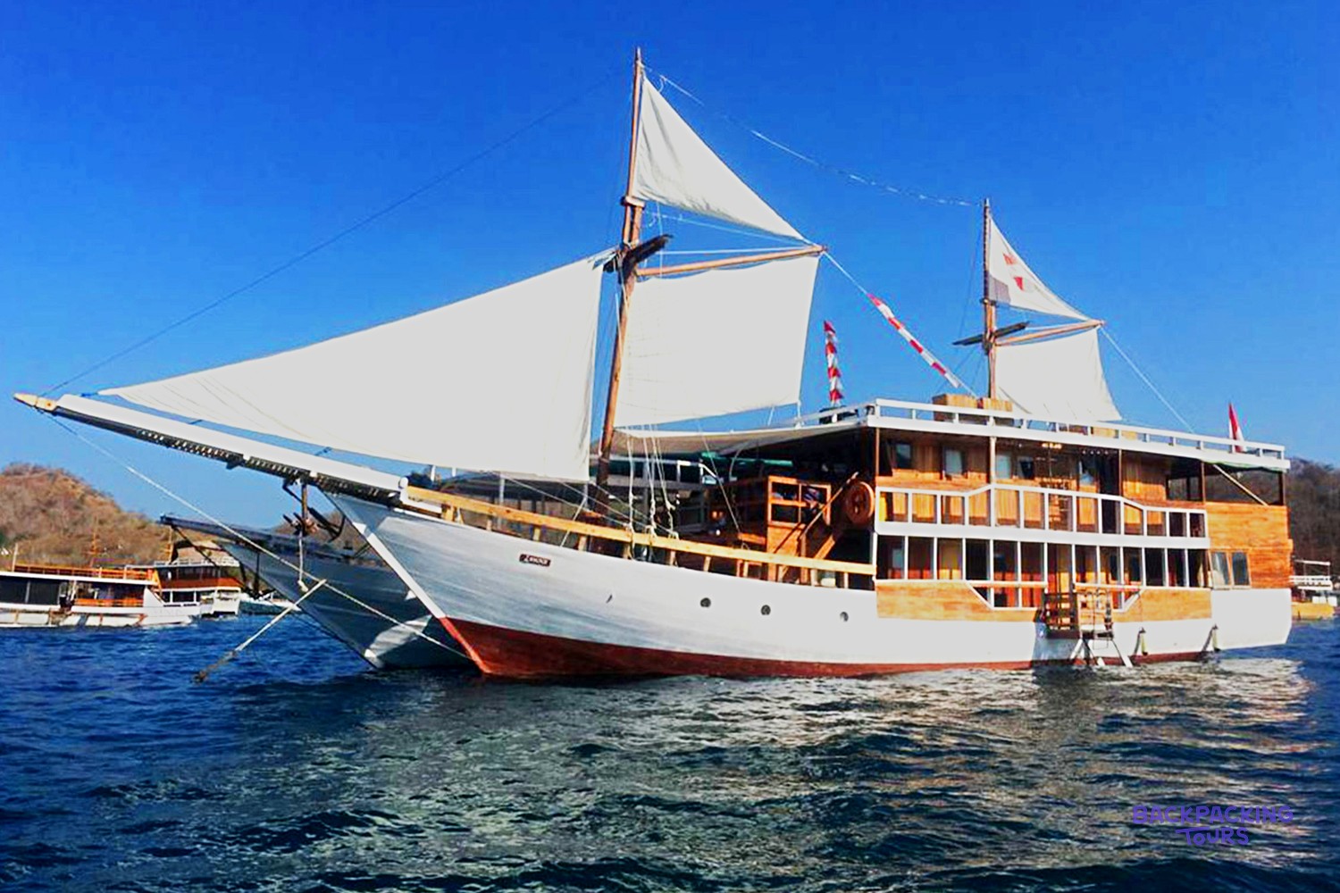 A large wooden boat with white sails floats on blue water under a clear sky, with hills in the background.