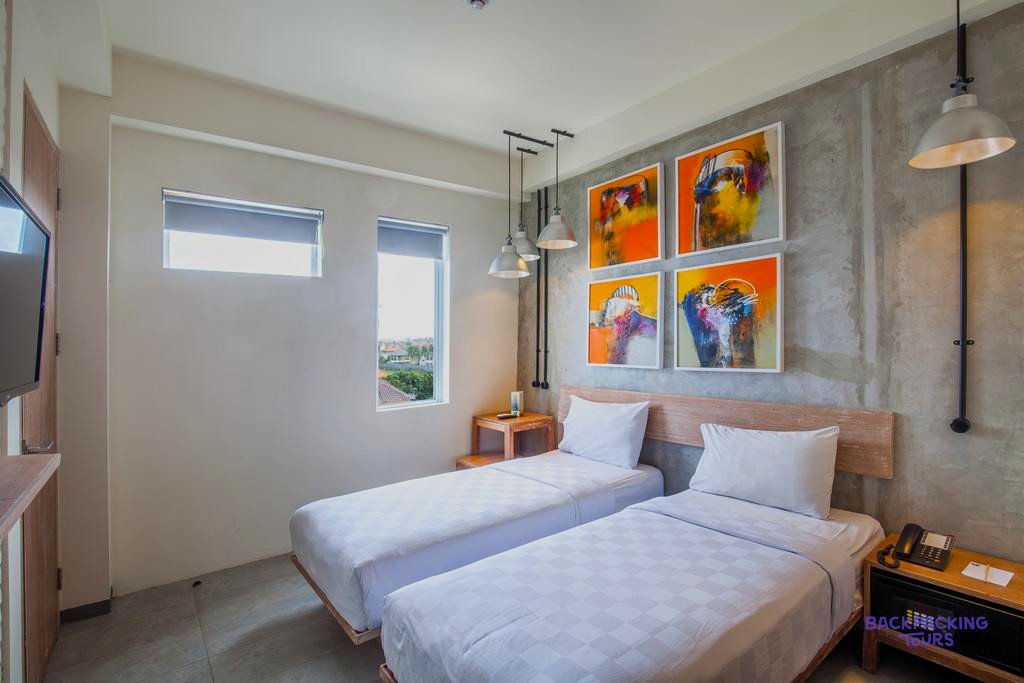A bright hotel room contains two single beds, white bedding, and colourful abstract artwork on the wall.