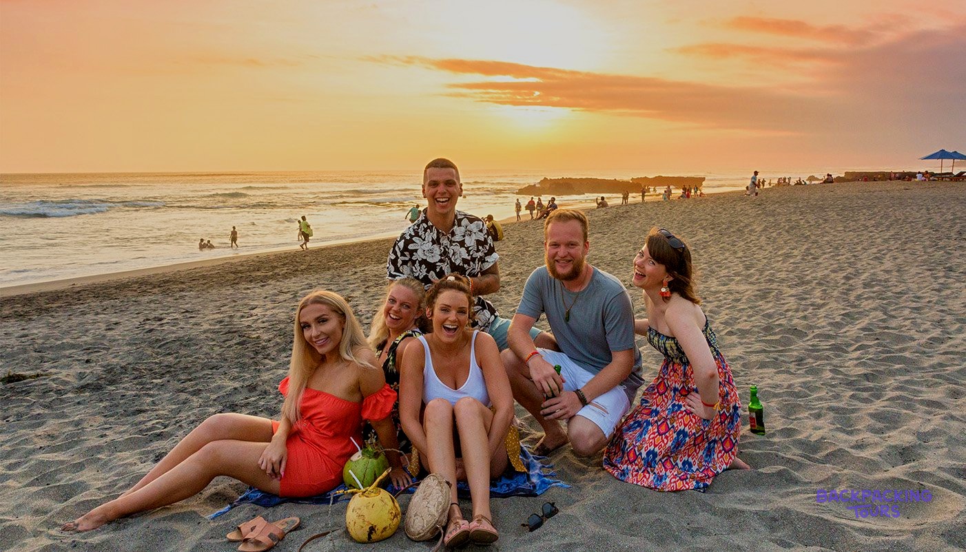 Six young people sit on a sandy beach with coconuts as the sun sets over the ocean.