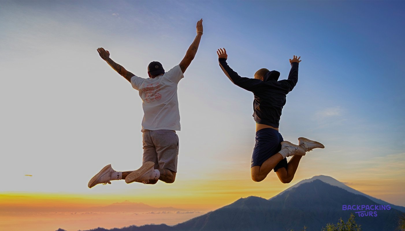Two people jump with raised arms against a colourful sunrise sky above mountain peaks.