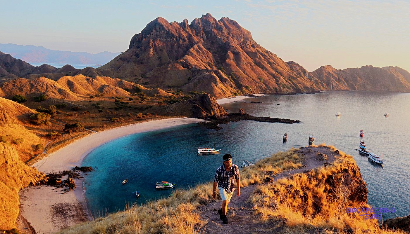 A person walks a dry grassy path above a bay with turquoise water, white sand, and distant mountains.