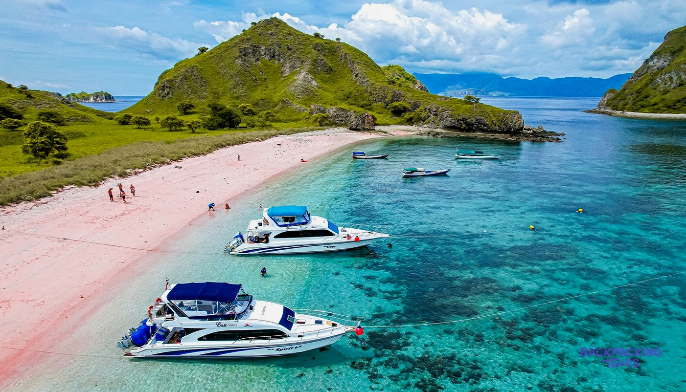 A pink sandy beach with clear turquoise water, boats, and green hills under a blue sky.