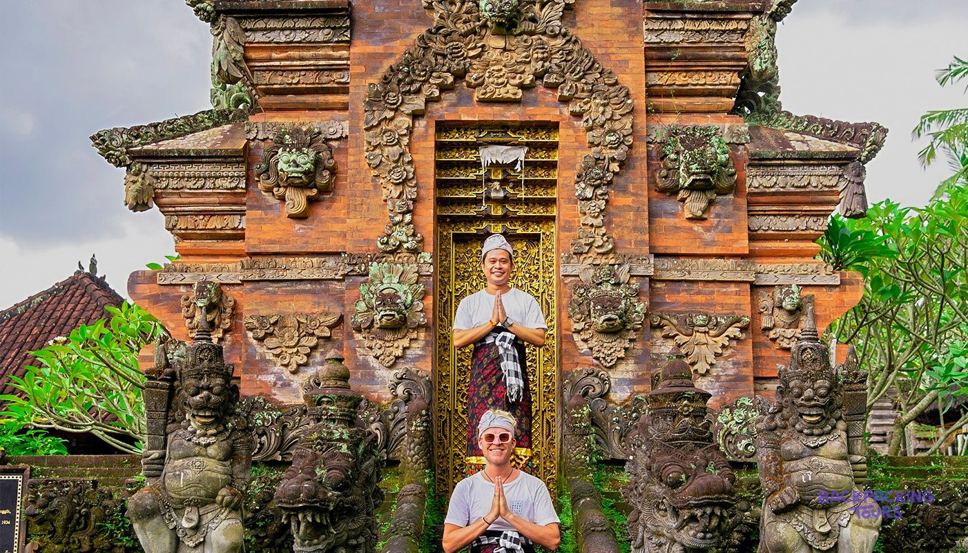 Two men in traditional dress pose with hands pressed together before an ornate brick temple gate.