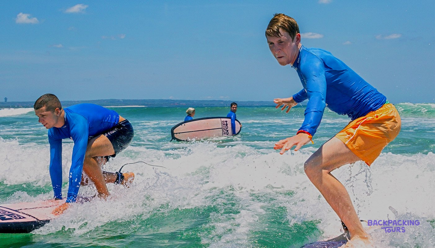 Two young men in blue tops are riding surfboards on green ocean waves.