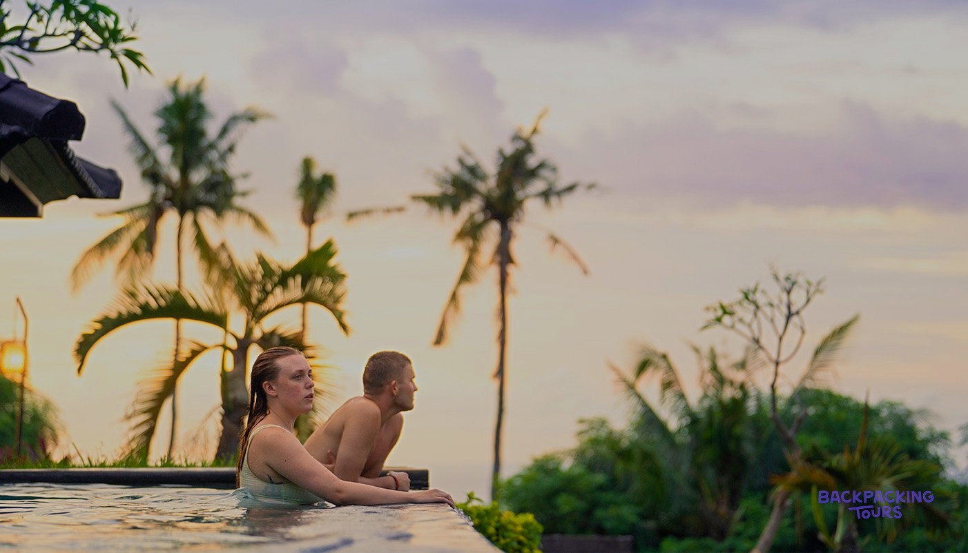 Two people relax in an infinity pool, looking out at palm trees against a sunset sky.