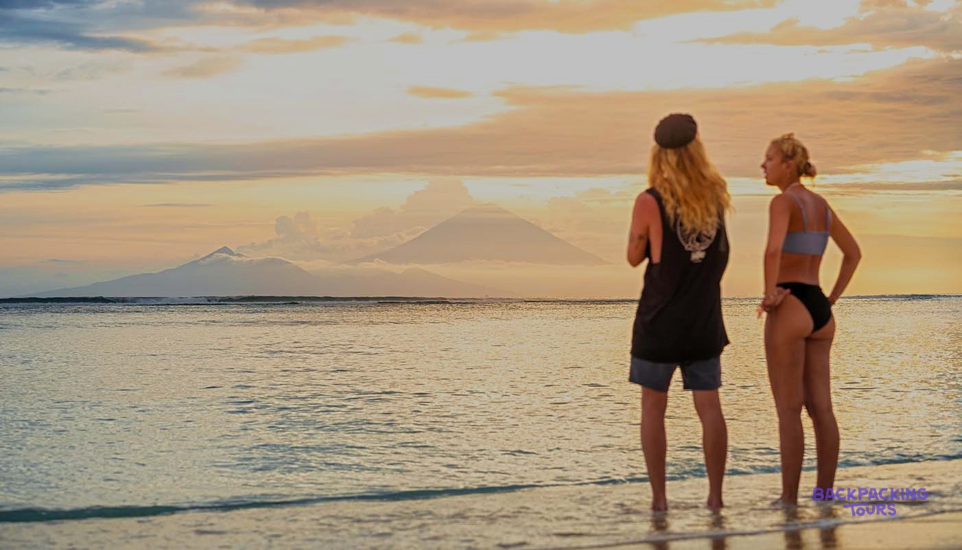 Two people stand in the shallow sea, looking at distant mountains under a colourful sunset sky.