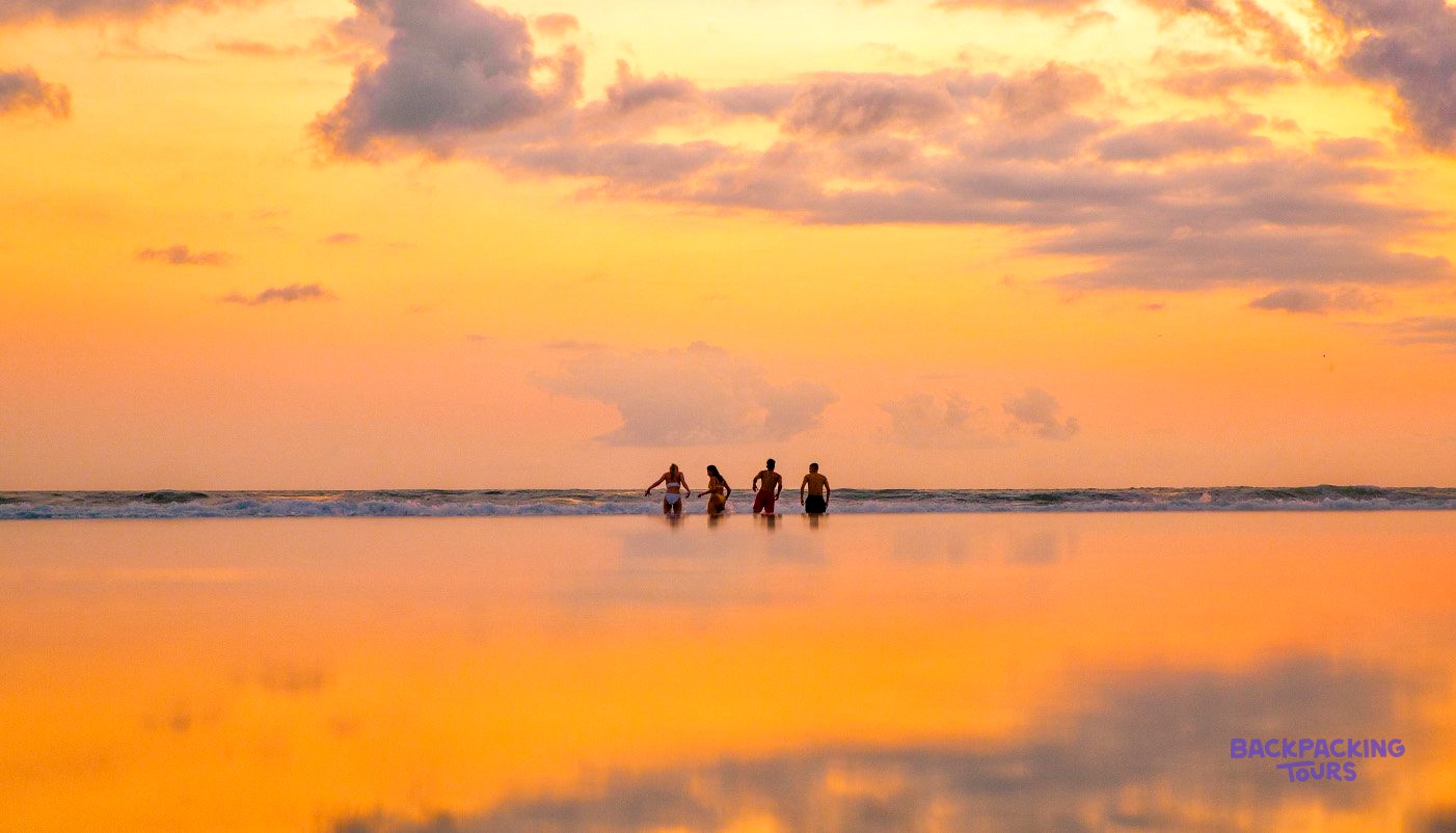 Four people wade in the ocean at sunset, with the sky and water bathed in orange and yellow hues.