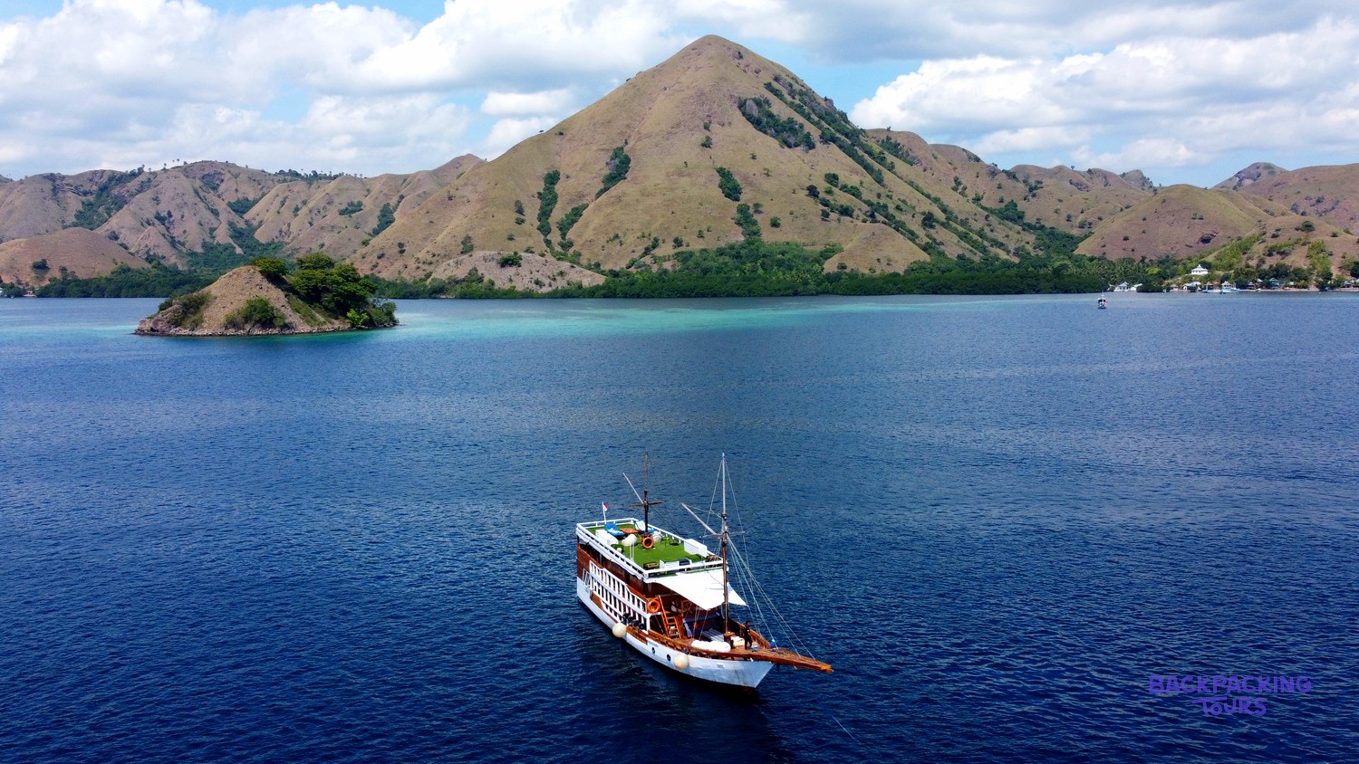 A large wooden sailing boat glides across deep blue water with dry, hilly islands in the background.