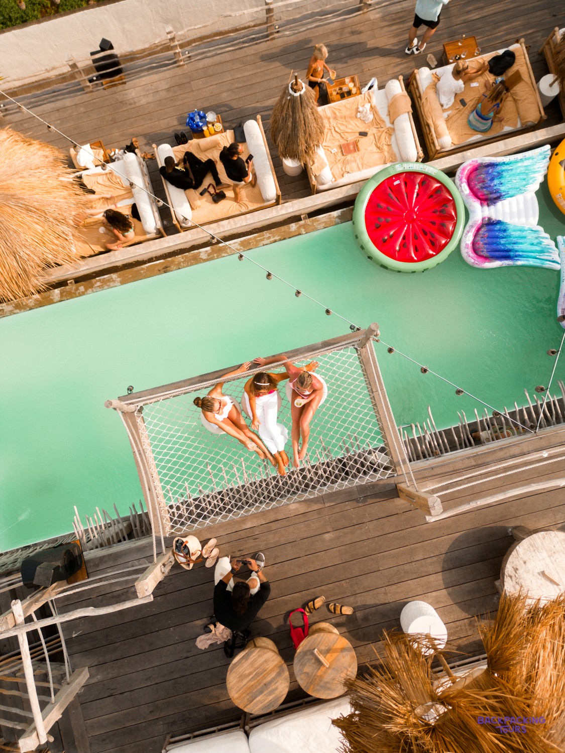 Three women relax in a net hammock over a turquoise swimming pool at a beach club.