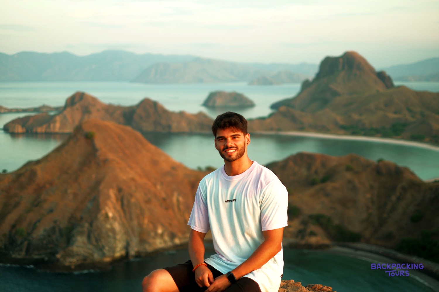 A young man sits on a rocky viewpoint overlooking many hilly islands in calm turquoise water.