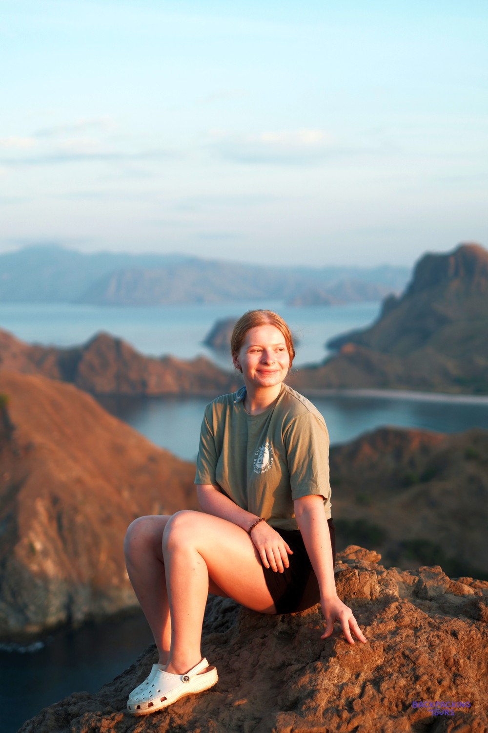 Young woman sits on a rocky viewpoint, overlooking a bay with many hilly islands.
