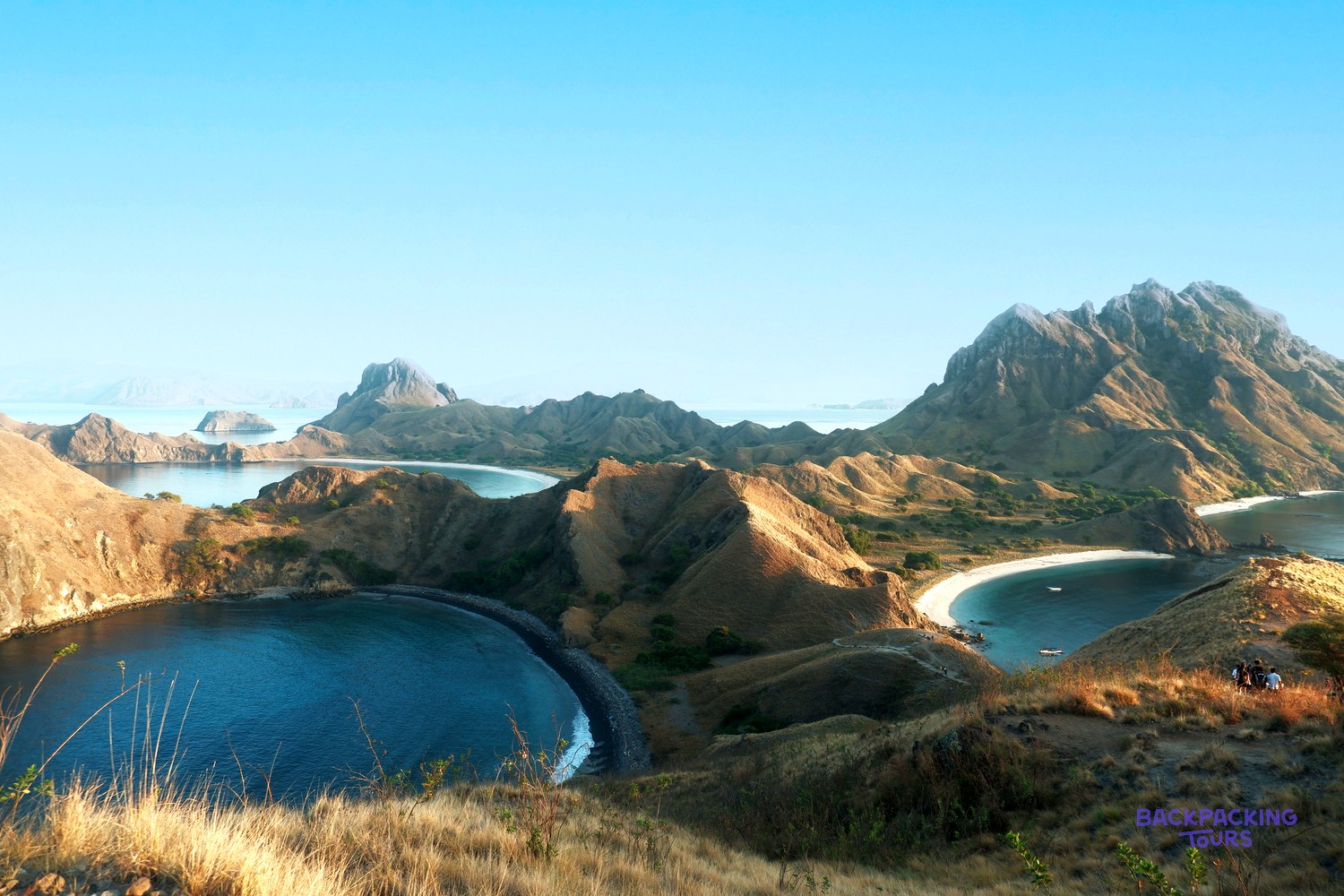 Rolling dry hills surround deep blue bays with white and black sand beaches under a clear sky.