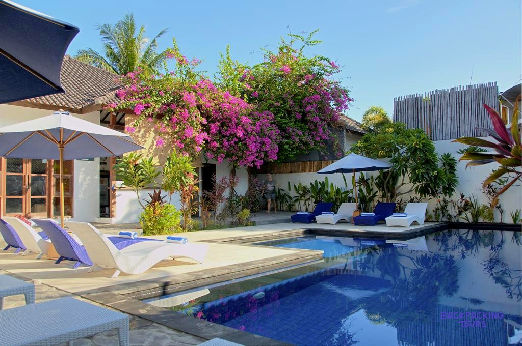 A sunny swimming pool area with sun loungers, blue umbrellas, and a building covered in vibrant pink flowers.