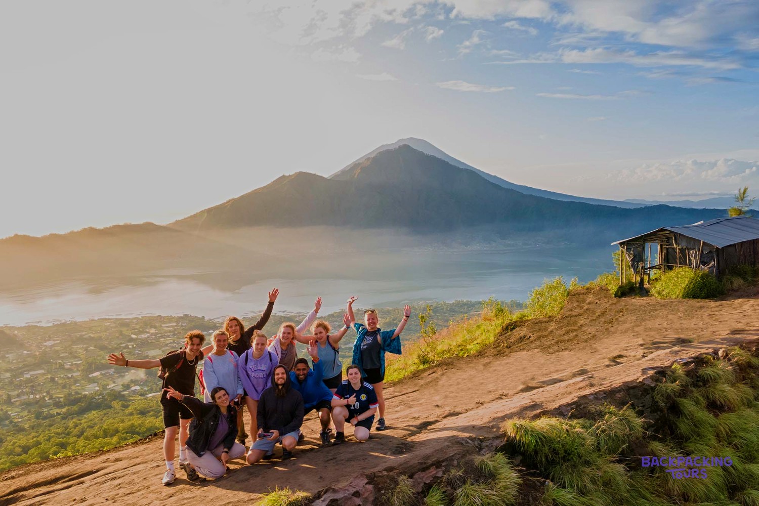 A group of people pose on a mountain slope at sunrise, overlooking a misty lake and distant volcanoes.