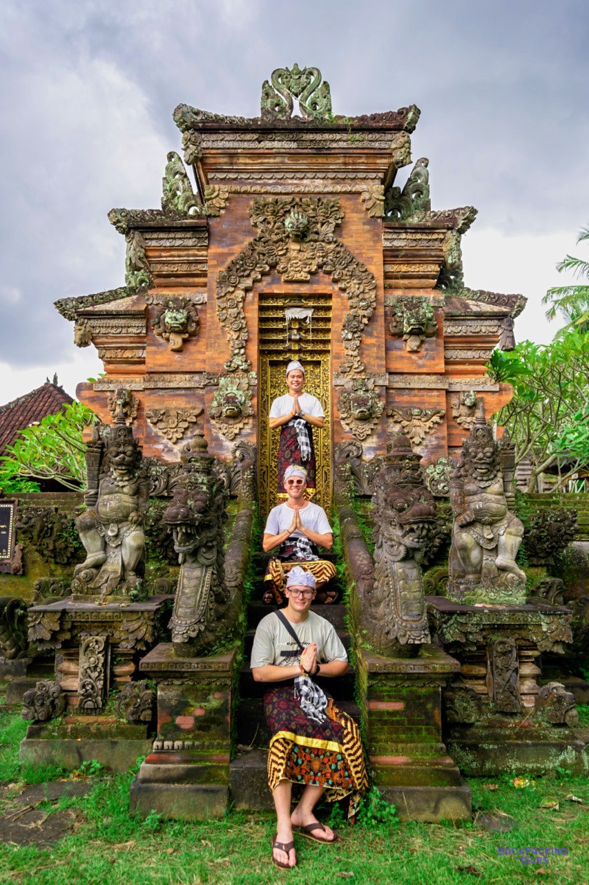 Three travellers in traditional sarongs pose with hands together on moss-covered temple steps.