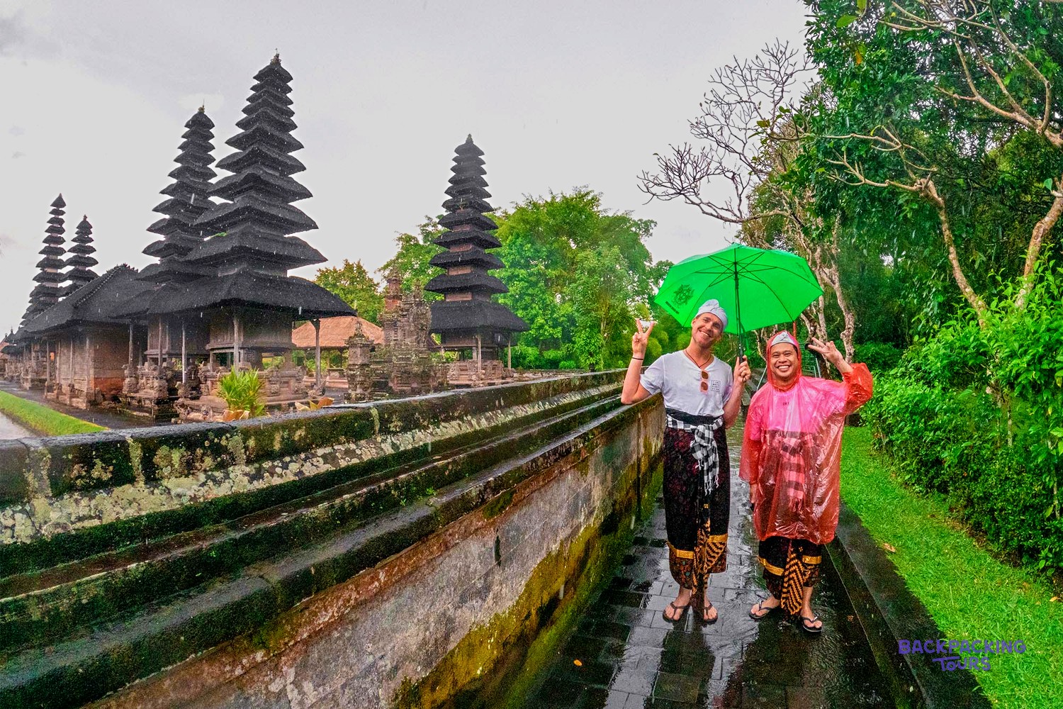 Two people pose with peace signs on a wet path, with multi-tiered temple structures and lush greenery behind them.