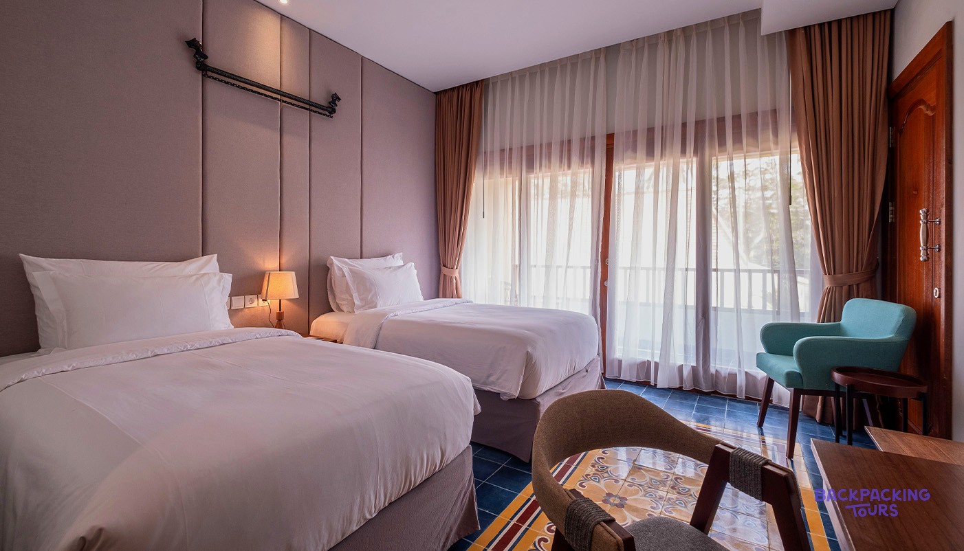 Hotel room with two single beds, a teal armchair, and patterned floor tiles next to a window.