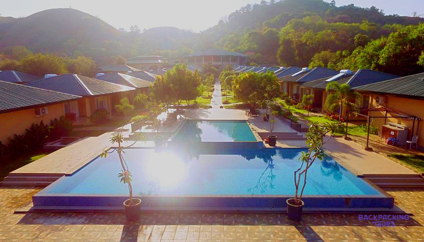 A large swimming pool reflecting sunlight, surrounded by rows of bungalows, with lush green hills in the background.