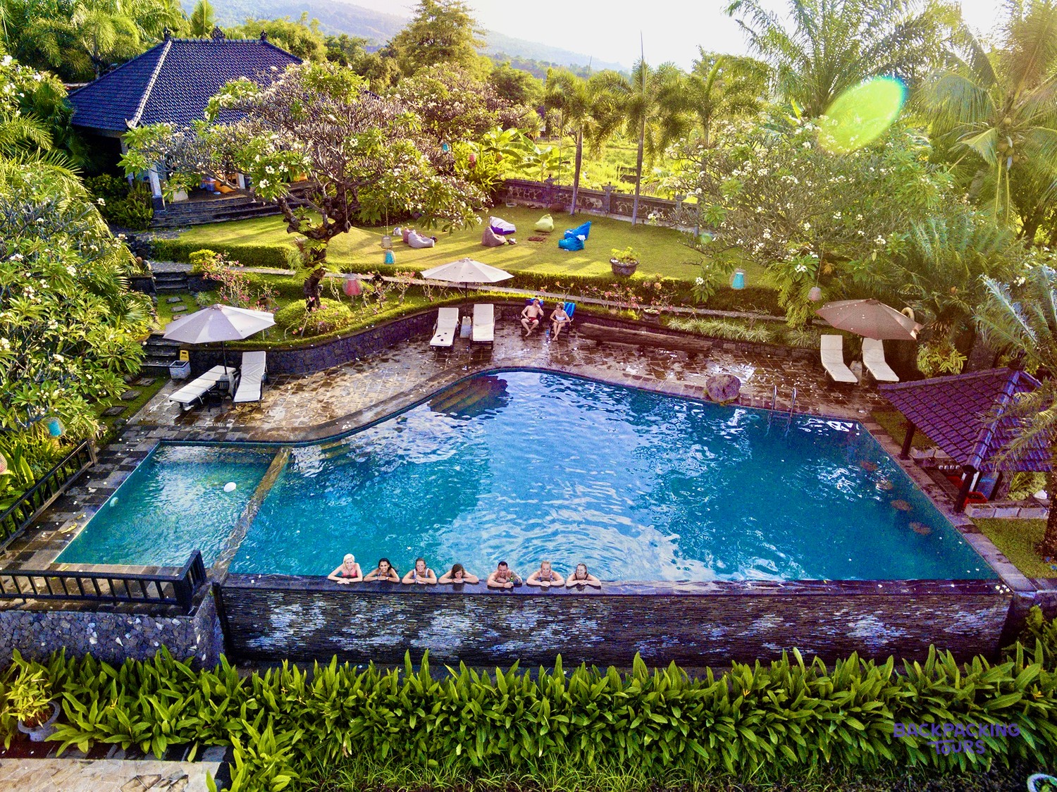 A private villa with a large swimming pool, people relaxing in the water, surrounded by lush gardens and palm trees.