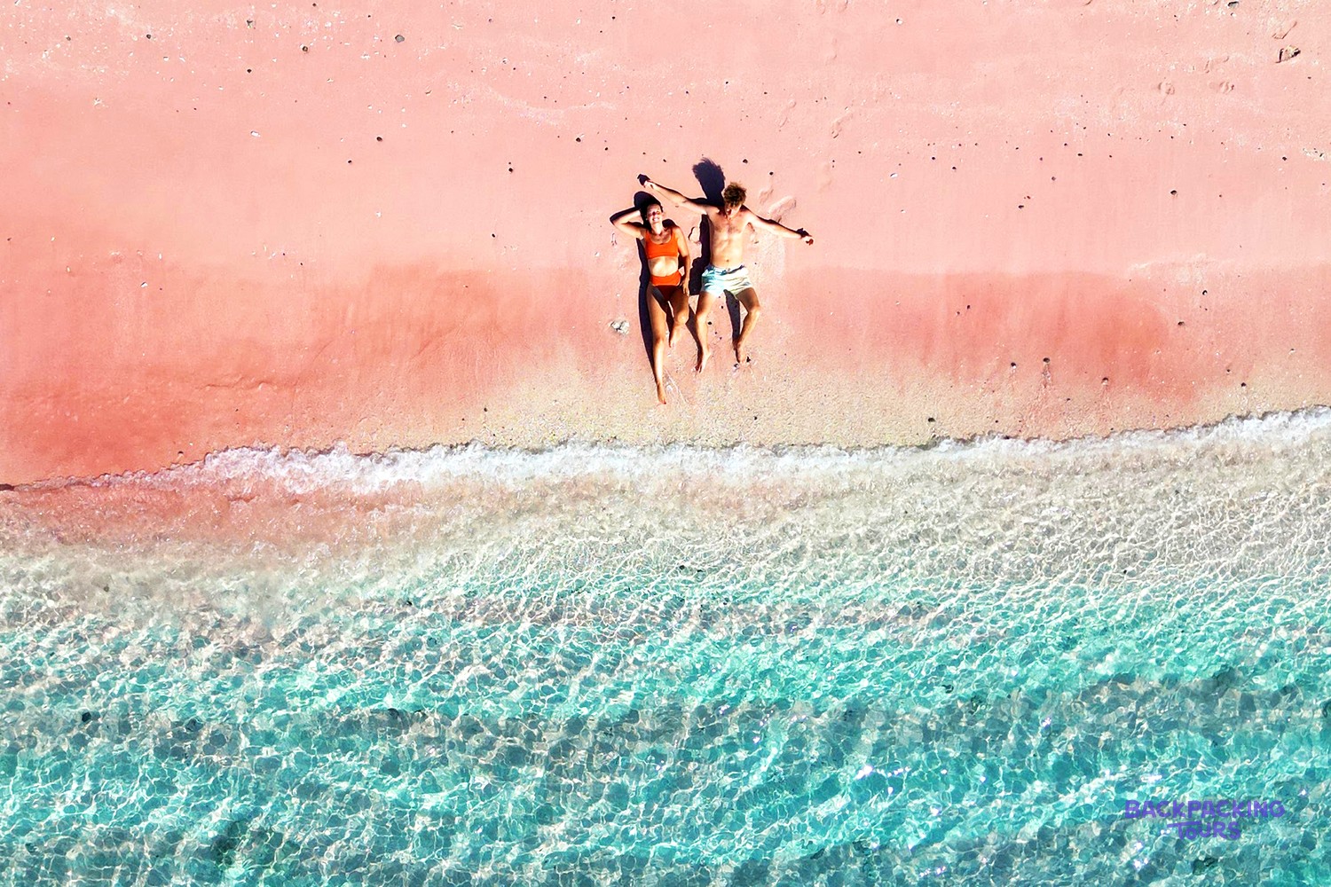 Two people lie on a pink sand beach beside clear turquoise water with gentle waves.