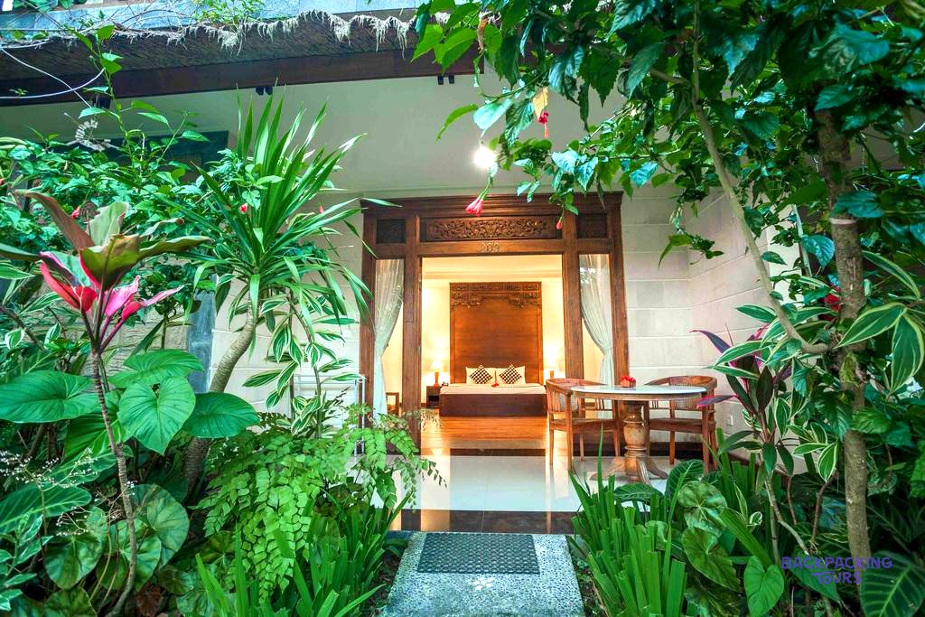 A wooden doorway leads to a bright bedroom with a bed, surrounded by lush tropical foliage.