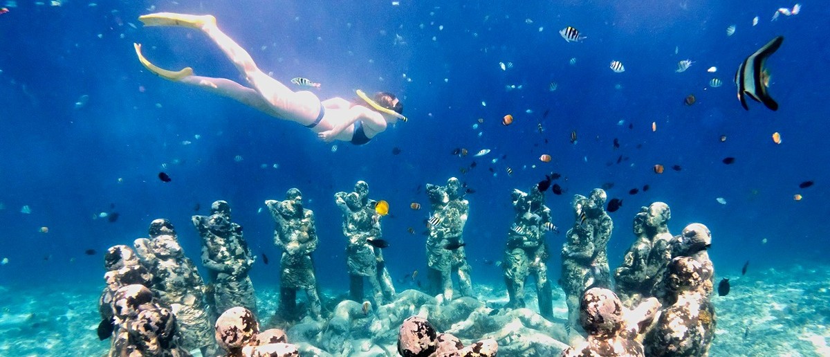 A snorkeller swims above a circle of human-like statues on the seabed, surrounded by colourful fish.
