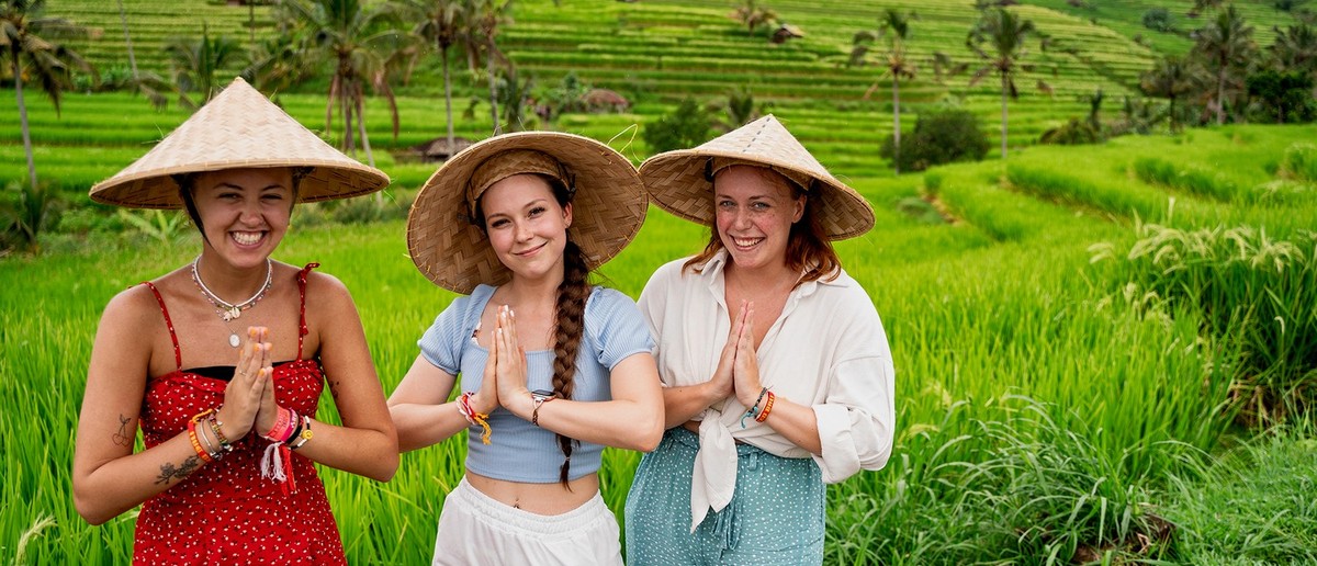 Three young women in straw hats stand with hands pressed together, surrounded by lush green rice terraces and palm trees.